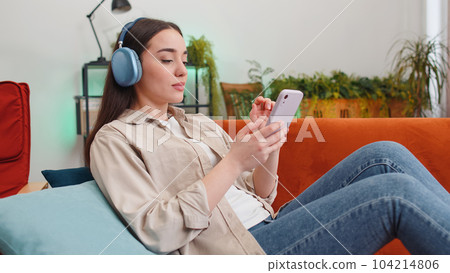 Happy young woman in wireless headphones relaxing lying on sofa at home listening favorite music Happy young woman in wireless headphones relaxing lying on sofa at home listening favorite music 104214806