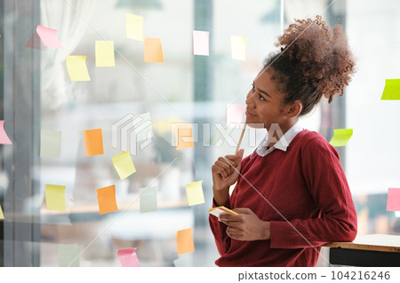 African American female employee write down on colorful sticky notes manage list, concentrated biracial woman work on startup brainstorm collaborate plan on stickers on glass wall 104216246