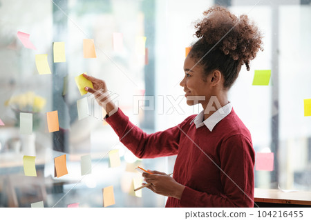 African American female employee write down on colorful sticky notes manage list, concentrated biracial woman work on startup brainstorm collaborate plan on stickers on glass wall 104216455