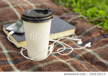 Cup of coffee, open book and smartphone with headphones on blanket in the park Cup of coffee, open book and smartphone with headphones on blanket in the park 104218213