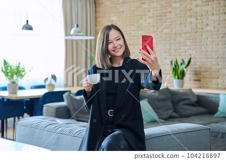 Young woman having video call sitting on sofa at home, using smartphone 104218897