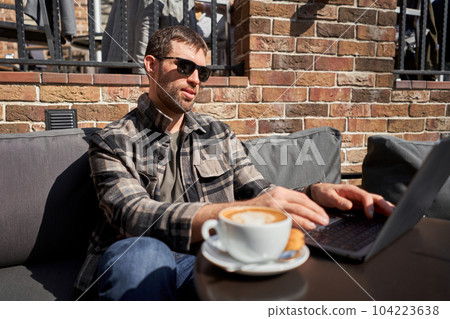 Portrait of a handsome successful smart man drinking coffee and looking at a laptop. A business man is having breakfast sitting on a beautiful terrace in a cafe. Reading, browsing the Internet 104223638