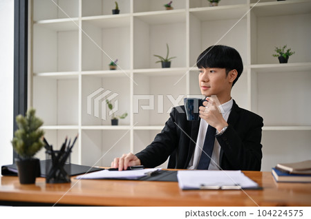 A handsome Asian male lawyer in a formal suit is sipping his morning coffee at his desk. 104224575