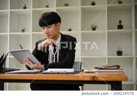A smart Asian businessman focuses on reading something on his tablet while sitting at his desk 104224578