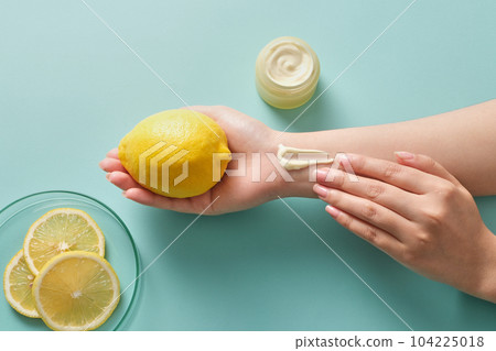 On a blue background, a fresh lemon on palm and the other hand applying cream texture. Mockup scene for advertising. Natural Vitamin C skincare beauty product concept. 104225018