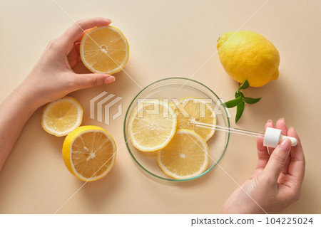 Female hand holding half a fresh lemon and a dropper with slices of lemon and green leaves decorated on beige background. Concept of natural cosmetics with lemon extract Female hand holding half a fresh lemon and a dropper with slices of lemon and green leaves decorated on beige background. Concept of natural cosmetics with lemon extract 104225024