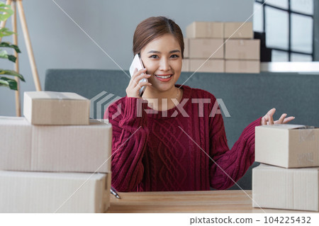 Cheerful small online business owner processing Internet store order for shipping, typing on laptop, speaking mobile phone, using gadgets for communication with clients 104225432