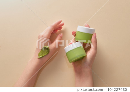 Two green jars is held by a woman hand model and a smear of cream texture on another hand. Mockup of skin care cosmetic jar 104227480