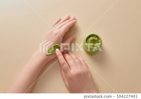 Top view of hand model applying green smear of creamy texture on her hand with a green jar beside. Light beige background 104227481