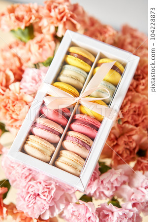 Dessert French Macarons in present box on a background of flowers. Sweet dessert on a flowers background. Still life with macaroni cakes and different flower. Delivery concept, coffee and flower shop 104228873