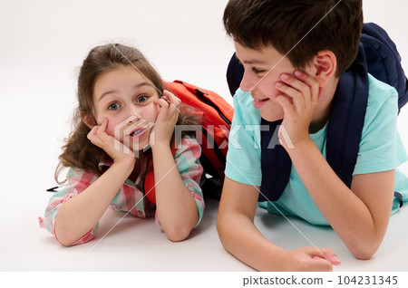 Two adorable smart school kids, a lovely first grader little child girl and her caring brother - a preteen schoolboy, lying on belly, smiling talking relaxing in recreation time over white background Two adorable smart school kids, a lovely first grader little child girl and her caring brother - a preteen schoolboy, lying on belly, smiling talking relaxing in recreation time over white background 104231345