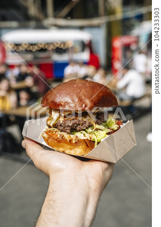 Street food burger on hand on hipster food market background. Grill and barbecue food Street food burger on hand on hipster food market background. Grill and barbecue food 104233303