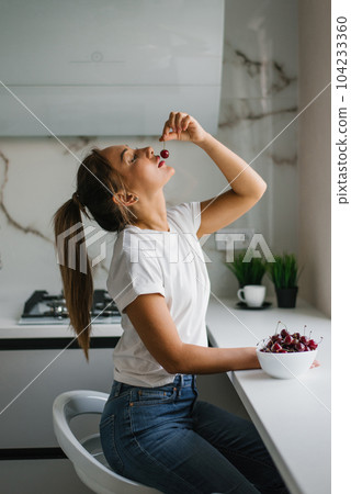 Attractive young woman eats fresh cherries, healthy berries sitting at home in the kitchen 104233360