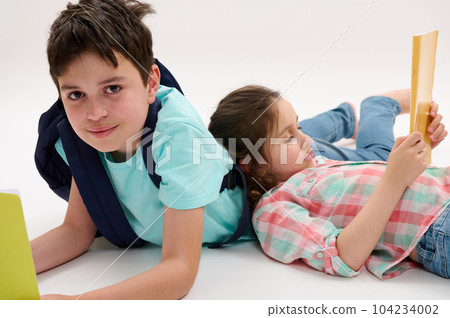 Portrait of Caucasian handsome preteen schoolboy with backpack and workbooks, lying near his younger sister, smiling looking at camera, isolated on white studio background. School kids. Back to school 104234002