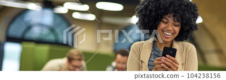Young happy mixed race female office worker looking at smartphone and feeling excited while standing in coworking space, colleagues on the background Young happy mixed race female office worker looking at smartphone and feeling excited while standing in coworking space, colleagues on the background 104238156
