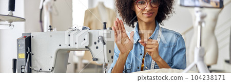 Smiling African-American seamstress shows black thread to camera sitting at workplace with sewing machine Smiling African-American seamstress shows black thread to camera sitting at workplace with sewing machine 104238171