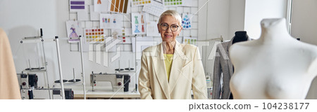 Mature woman with glasses stands at table with clothes drawings Mature woman with glasses stands at table with clothes drawings 104238177