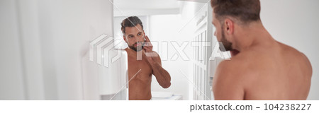 Positive man applies moisturizing cream onto face skin near large mirror in bathroom Positive man applies moisturizing cream onto face skin near large mirror in bathroom 104238227