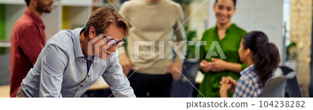 Happy businessman standing in the modern coworking space, looking at laptop screen and smiling while his young multiracial colleagues communicating on the background Happy businessman standing in the modern coworking space, looking at laptop screen and smiling while his young multiracial colleagues communicating on the background 104238282