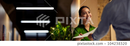 Young cheerful asian woman giving some documents to her colleague and smiling while standing together in the modern office Young cheerful asian woman giving some documents to her colleague and smiling while standing together in the modern office 104238320