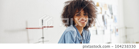 African-American seamstress with smartphone rests leaning onto cutting table in workshop 104238330
