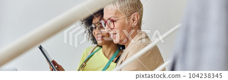 Middle aged woman with tablet sits with African-American colleague working on laptop on staircase 104238345