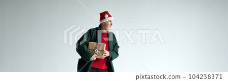Full length shot of cool middle aged man with Christmas hat wearing casual outfit looking away, holding gift box while posing isolated over light gray background 104238371