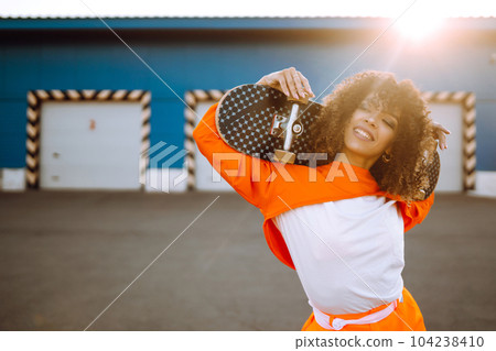 Beautiful African American woman posing with a skateboard. Hipster woman with curly hair with skateboard at sunset. Beautiful African American woman posing with a skateboard. Hipster woman with curly hair with skateboard at sunset. 104238410