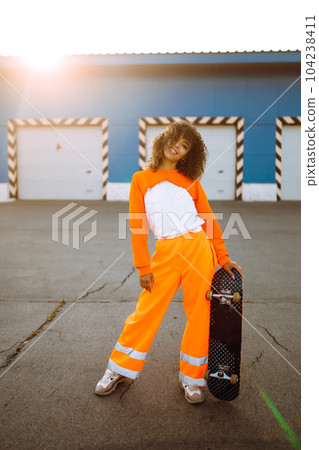 Beautiful African American woman posing with a skateboard. Hipster woman with curly hair with skateboard at sunset. Beautiful African American woman posing with a skateboard. Hipster woman with curly hair with skateboard at sunset. 104238411
