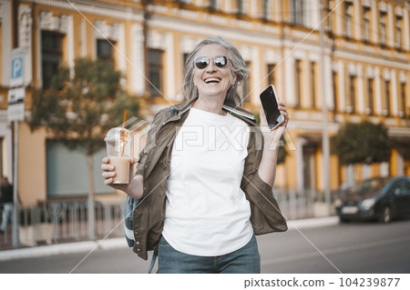 Happy senior old woman smiling outdoors. She speaking on phone, engaging in modern communication, while enjoying refreshing drink from transparent plastic cup. Mature lady in moment of joy. High 104239877