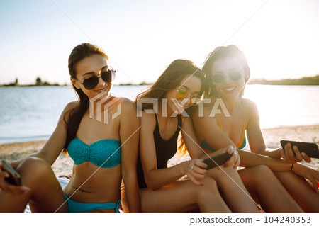 Three beautiful girls using mobile smart phone at the beach. Girls sunbathing on the beach. 104240353