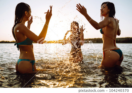 Three young girls splashing water in the sea. Happy friends having fun in the waves at sunset time. 104240370