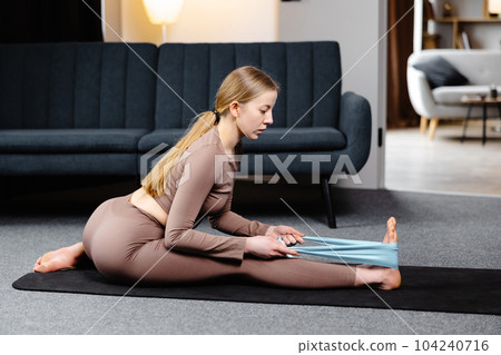 Young yogi woman practicing yoga concept, doing exercise stretching legs with elastic band, wearing sportswear bra and pants on the floor in living room 104240716