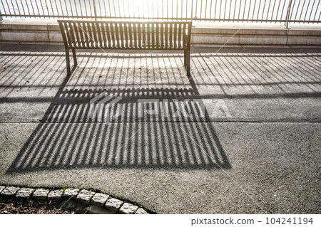 Morning sun cast lines of shadows on asphalt pavement through fence and empty bench by the river Morning sun cast lines of shadows on asphalt pavement through fence and empty bench by the river 104241194