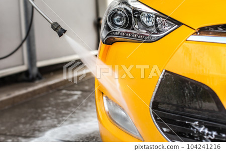 Front bumper and fog light of yellow car washed in self serve carwash; detail on jet water from nozzle spraying on glossy surface. Front bumper and fog light of yellow car washed in self serve carwash; detail on jet water from nozzle spraying on glossy surface. 104241216