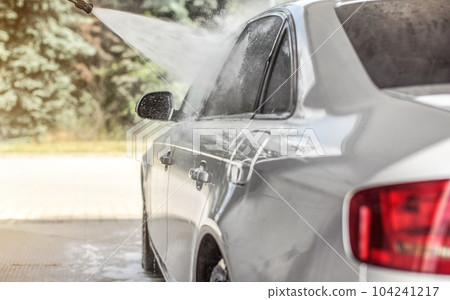 Silver car washed in self service carwash, jet water spraying from nozzle to side window, sun lit trees in background. Silver car washed in self service carwash, jet water spraying from nozzle to side window, sun lit trees in background. 104241217