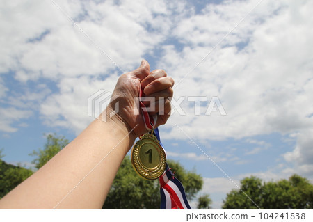 Blurred of woman hands raised and holding gold medals with Thai ribbon against blue sky background to show success in sport or business, Winners success award concept. Blurred of woman hands raised and holding gold medals with Thai ribbon against blue sky background to show success in sport or business, Winners success award concept. 104241838