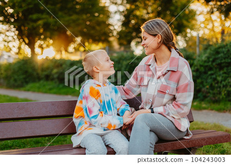 mother and son sit on a park bench in the rays of the setting sun. the concept of a family. Mother's Day. beautiful girl (mother) with a boy (son) in the park in the park are sitting on a bench at mother and son sit on a park bench in the rays of the setting sun. the concept of a family. Mother's Day. beautiful girl (mother) with a boy (son) in the park in the park are sitting on a bench at 104243003