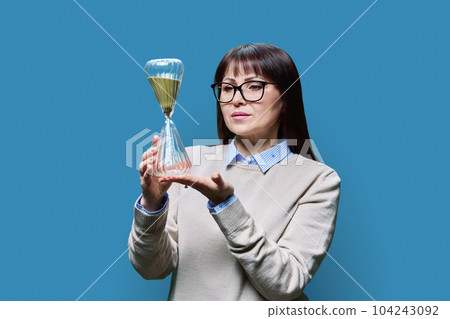 Middle aged woman with hourglass on blue studio background Middle aged woman with hourglass on blue studio background 104243092