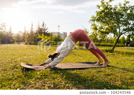 Side view of healthy female yoga practitioner preforming Downward Facing Dog Pose Adho Mukha Svansana at city park on sunny summer day. Concept of outdoors yoga, fitness, active and sporty lifestyle. 104243546