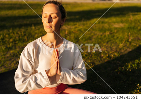 Portrait of relaxed young woman practicing yoga performing namaste pose with closed eyes at city park on sunny summer sunset. Pretty calm female sitting lotus position on yoga mat outdoors alone. Portrait of relaxed young woman practicing yoga performing namaste pose with closed eyes at city park on sunny summer sunset. Pretty calm female sitting lotus position on yoga mat outdoors alone. 104243551