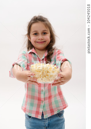 Happy cheery Caucasian little girl in casual clothes, holding out at camera a bowl with salted popcorn, ready to watch cartoon or movie, smiling on white background. People. Kids. Food. Entertainment 104245018