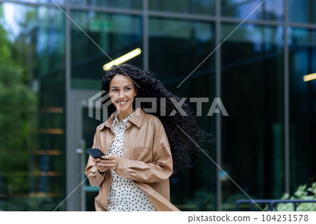 Young beautiful hispanic woman walking in the city, business woman holding phone in hands using smartphone app, woman smiling contentedly and happy outside office building with curly hair 104251578