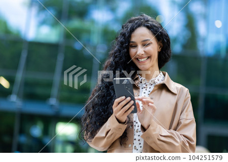 Young beautiful hispanic woman walking in the city, business woman holding phone in hands using smartphone app, woman smiling contentedly and happy outside office building with curly hair 104251579