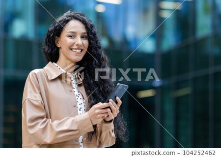 Portrait of a successful and satisfied Latin American business woman, female boss smiling and looking at the camera, holding a smartphone, using an online application, dialing a message and a call. 104252462