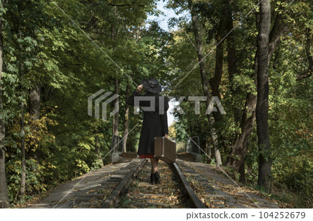 Aristocrat Lady in black coat and hat of 40s style with retro valise in hands walks along rails going through park. Back view Aristocrat Lady in black coat and hat of 40s style with retro valise in hands walks along rails going through park. Back view 104252679