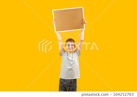 Happy boy with glasses holds cork gloss above head with space for text. Schoolboy holds blank board on yellow background. Copy space. Mock up. 104252763