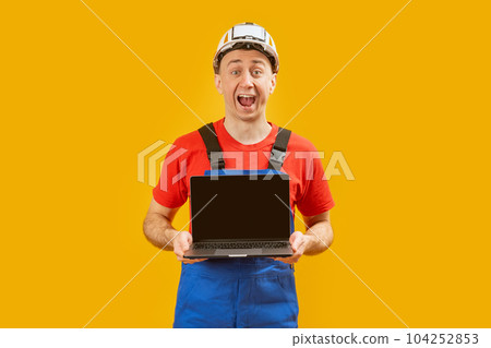 Handsome male foreman engineer holding laptop with blank screen on the yellow background. Copy space, mock up. 104252853