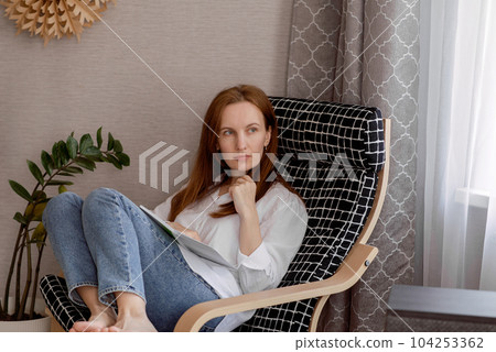 A young smiling woman, a happy confident woman in her 30's, holding a BLOCKET OR BOOK, sitting in a chair at home, working, looking at the camera. High quality photo 104253362