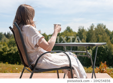 Woman relaxing on terrace, sitting in chair at table, drinking tea, enjoying view on nature landscape on summer holiday 104254856